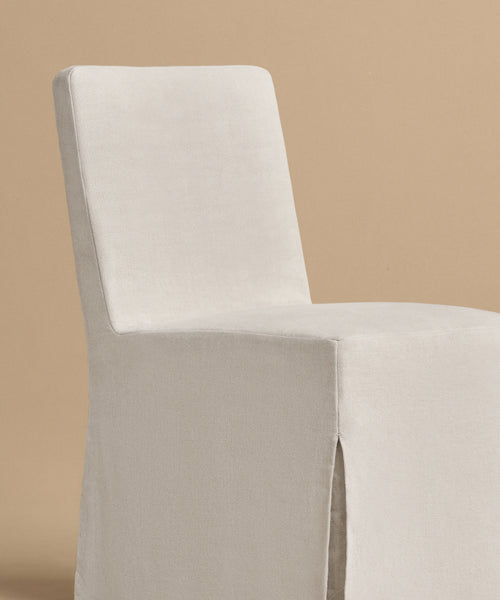 The Sunset Dining Chair showcases a modern, minimalist California design with a high rectangular back and light beige linen upholstery. Fabric skirt covers the legs. Photographed against a plain tan background.