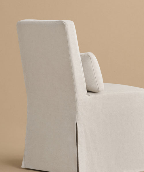 The Sunset Dining Chair, featuring beige linen upholstery, a straight backrest, and a matching rectangular cushion inspired by California design, stands against a plain light brown background.