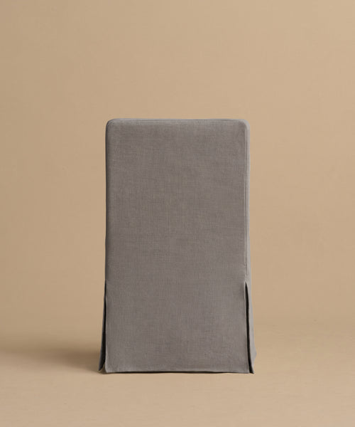 The Sunset Dining Chair, with a straight back and gray fitted linen slipcover with side slits, stands against a plain beige backdrop, showcasing effortless California design.