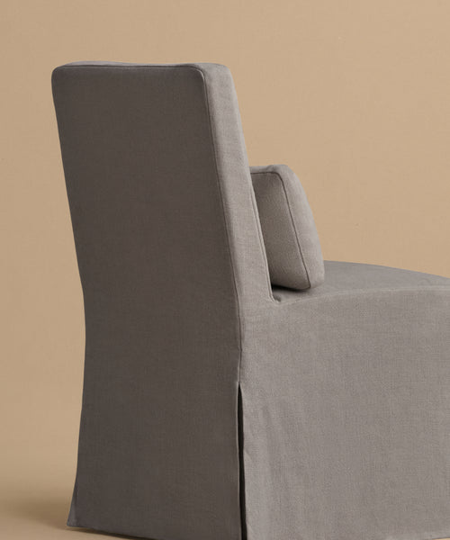 The Sunset Dining Chair, a modern armless design with a high back and matching lumbar cushion in plain gray linen, is showcased against a neutral beige background, embodying classic California style.