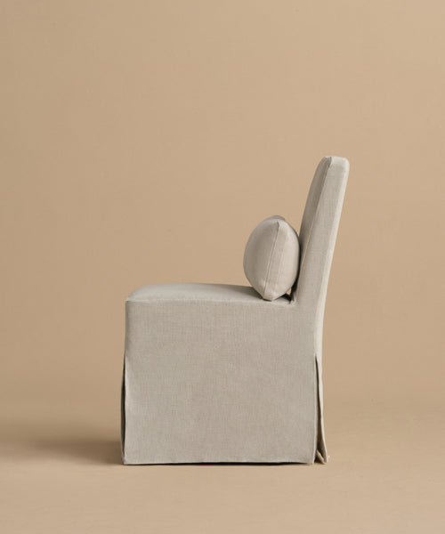 The Sunset Dining Chair, shown in profile against a beige background, features a light beige removable slipcover, skirted base, and round cushion on the backrest for added elegance and easy maintenance.
