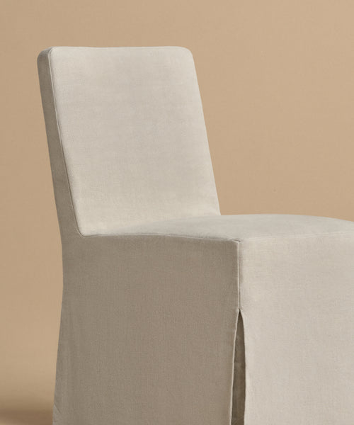 The Sunset Dining Chair, modern and armless with a high backrest, is fully wrapped in light beige fabric. It features a removable slipcover, a subtle front slit at the bottom, and is set against a plain beige background.