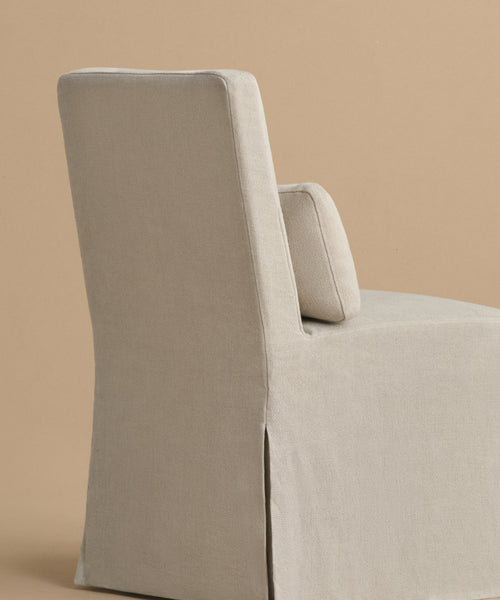 The Sunset Dining Chair is a modern, armless seat with a high back and light beige linen upholstery, shown angled left on a solid tan background.