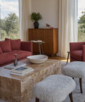 A modern living room with red armchairs and sofa, a Sur Rectangle Coffee Table topped with books and decor, two fuzzy white stools, tall white curtains, and a wooden cabinet by a large window overlooking scenic views.
