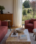 A cozy living room with pink armchairs, a Sur Rectangle Coffee Table topped with books and decor, a wooden side table, cabinet, and large window with sheer curtains overlooking a sunlit green landscape.