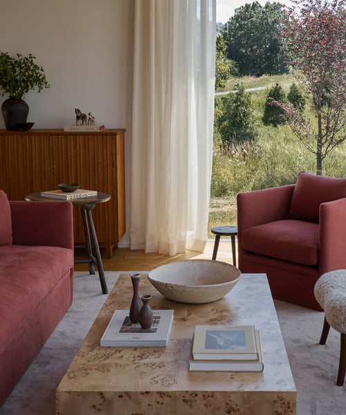 A cozy living room with pink armchairs, a Sur Rectangle Coffee Table topped with books and decor, a wooden side table, cabinet, and large window with sheer curtains overlooking a sunlit green landscape.