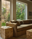 An upholstered sofa with matching cushions in a living room setting, positioned between large windows showing a green garden outside. Two burlwood side tables sit in front of the sofa.