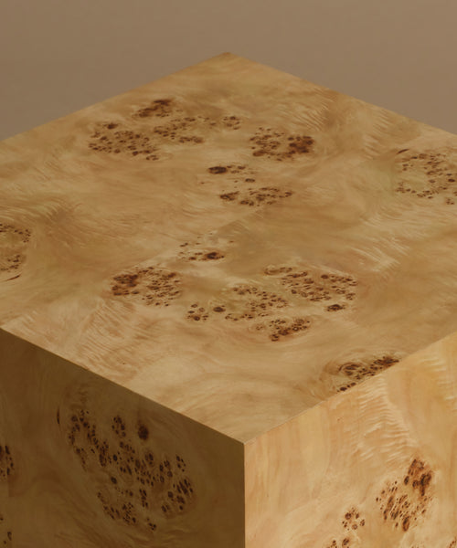A close up of a square burl wood side table standing upright on a beige surface against a solid beige background.