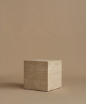 A square travertine side table, standing upright on a matching beige surface against a solid beige background.