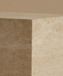A close up of a square travertine side table, standing upright on a matching beige surface against a solid beige background.