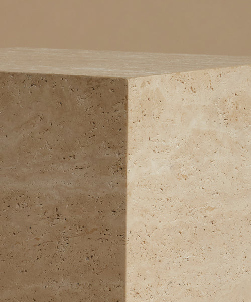 A close up of a square travertine side table, standing upright on a matching beige surface against a solid beige background.