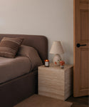 A styled bedroom corner featuring a dark brown upholstered bed, a light beige stone-look nightstand, a cream table lamp, a small candle, and a wooden door.