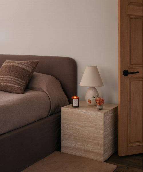 A styled bedroom corner featuring a dark brown upholstered bed, a light beige stone-look nightstand, a cream table lamp, a small candle, and a wooden door.