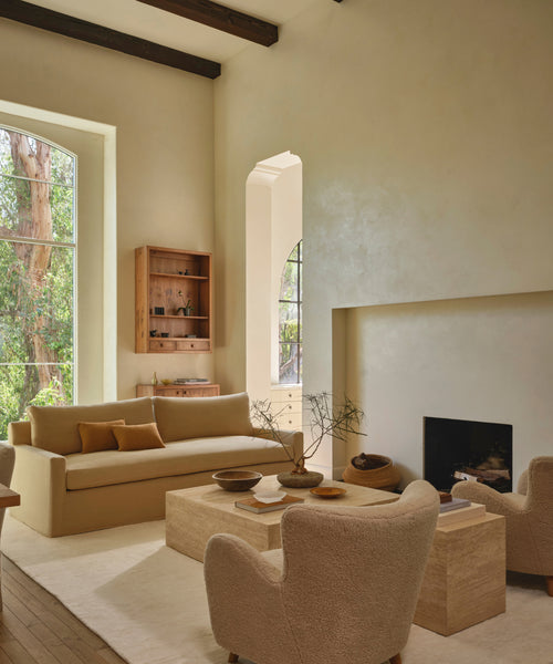A warm, modern living room featuring a beige sofa, textured armchairs, a low travertine coffee table, and a large white rug, set against a backdrop of textured plaster walls and a fireplace.