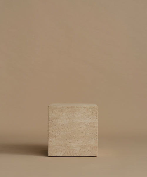A square travertine side table, standing upright on a matching beige surface against a solid beige background.