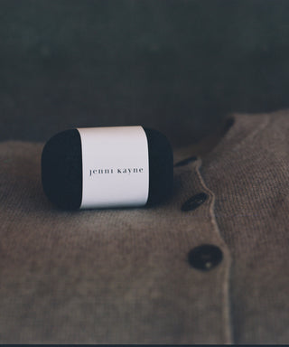 A black lint roller with a white Jenni Kayne label rests on a beige cardigan, perfectly paired with the Free Gift: Sweater Stone—made from natural pumice to remove pilling and keep your knits fresh.