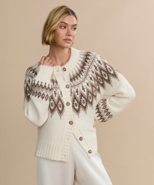 A woman with short blonde hair wears the Thatcher Crewneck Cardigan, an alpaca blend in cream with a brown geometric pattern and large buttons, paired with white pants against a beige background.