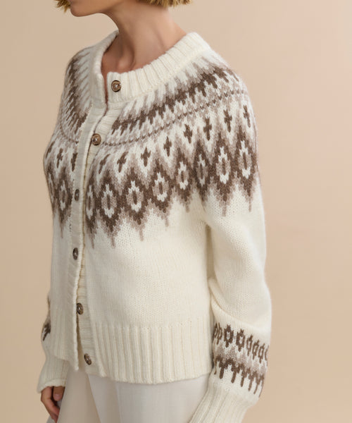 A person wears the Thatcher Crewneck Cardigan, a cream alpaca blend knit with a brown and white Fair Isle pattern on the shoulders and upper sleeves, ribbed collar, and brown buttons, standing against a beige background.