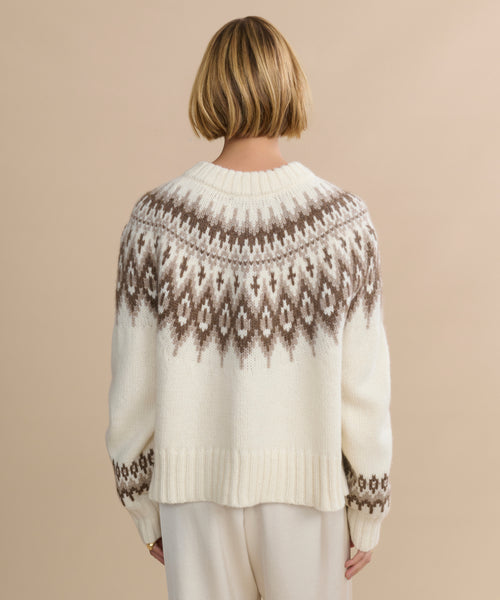 A person with short blonde hair is seen from behind wearing the Thatcher Crewneck Cardigan, a white alpaca blend sweater featuring a brown and gray geometric Fair Isle pattern. The background is light beige.