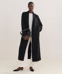 A woman wears the long black Theodore Coat in a plush wool-alpaca blend over a light top and wide white pants, styled with black pointed shoes and a black crossbody bag against a plain, neutral background.