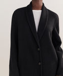 Someone is shown from the chest down wearing the Theodore Coat, a black, long-sleeve, buttoned cardigan with a shawl collar made from a wool-alpaca blend, layered over a white top against a neutral background.