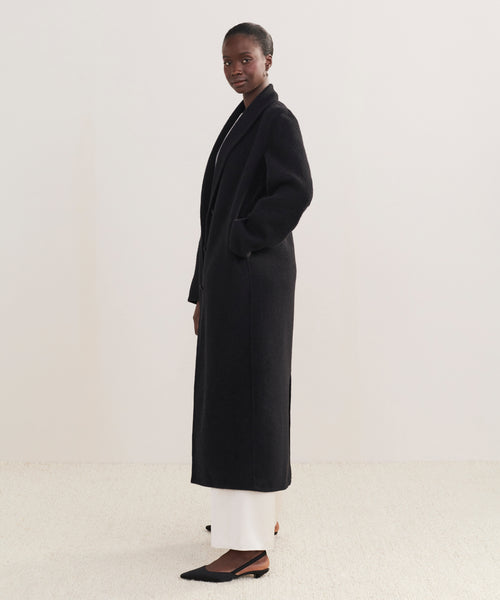 A woman with short hair stands in profile against a light background, wearing the Theodore Coat—a long black wool-alpaca blend coat—with white pants and black pointed-toe flats. Her hands are in the pockets of the coat.