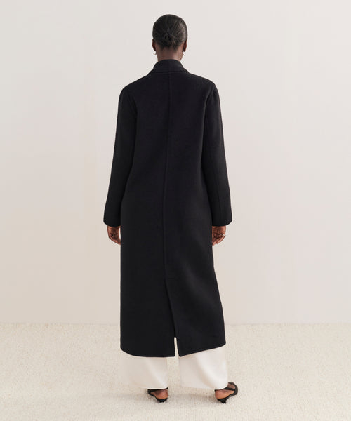 A person stands facing away in the mid-calf Theodore Coat, crafted from a wool-alpaca blend, paired with white wide-leg pants and black flat shoes against a plain light background.