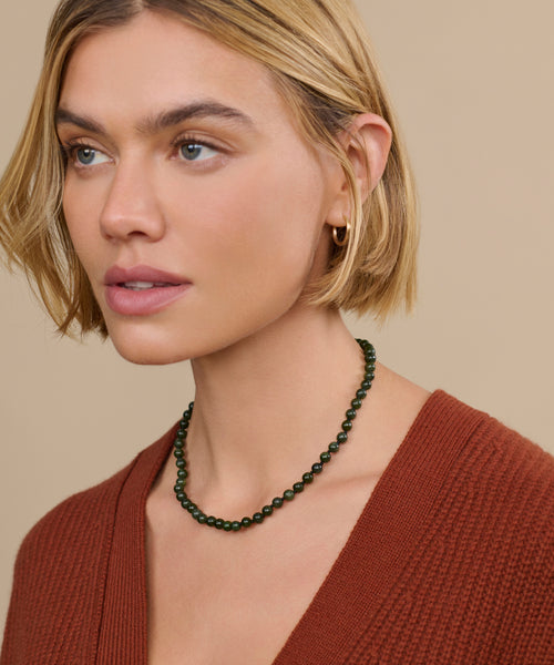 A woman with short blonde hair wears a rust-colored sweater, a small gold hoop earring, and the Tiny Boule Collar—a dark green heirloom-quality beaded necklace—while looking slightly to the side against a beige background.
