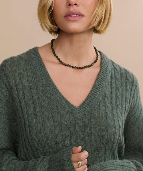 A woman with short blonde hair, partially visible against a neutral background, wears a textured green V-neck sweater and the Tiny Boule Collar—a piece of modernist jewelry—while holding her sweater with one hand.