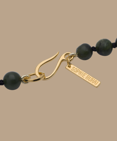Close-up of the gold clasp with a rectangular SOPHIE BUHAI tag on the Tiny Boule Collar, a black and dark green beaded necklace, shown against a beige background.
