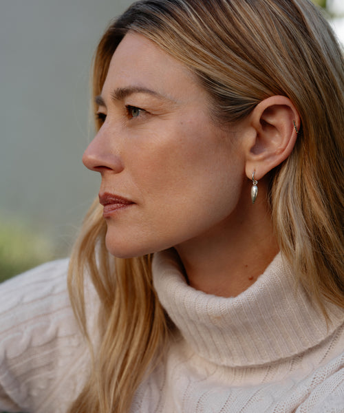 A woman with long blonde hair wears a cream-colored turtleneck sweater and Tiny Spear Hoops silver earrings, gazing thoughtfully to the left in a softly lit outdoor setting.