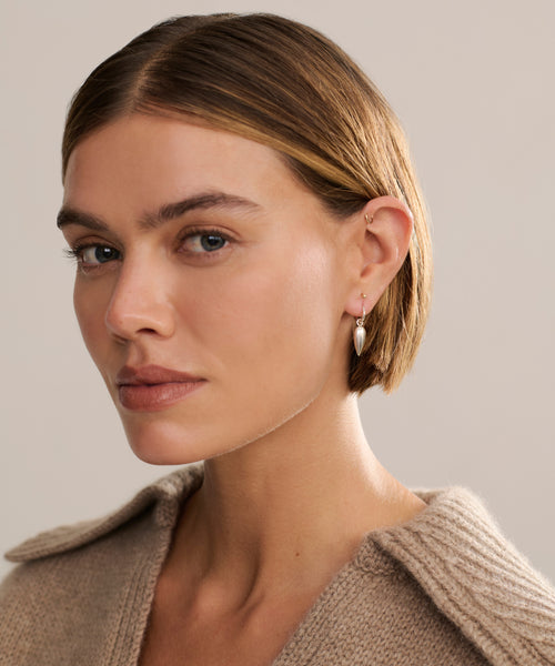 A woman with short, light brown hair and blue eyes wears a beige knit sweater and the elegant Tiny Spear Hoops, gazing calmly at the camera against a neutral background.