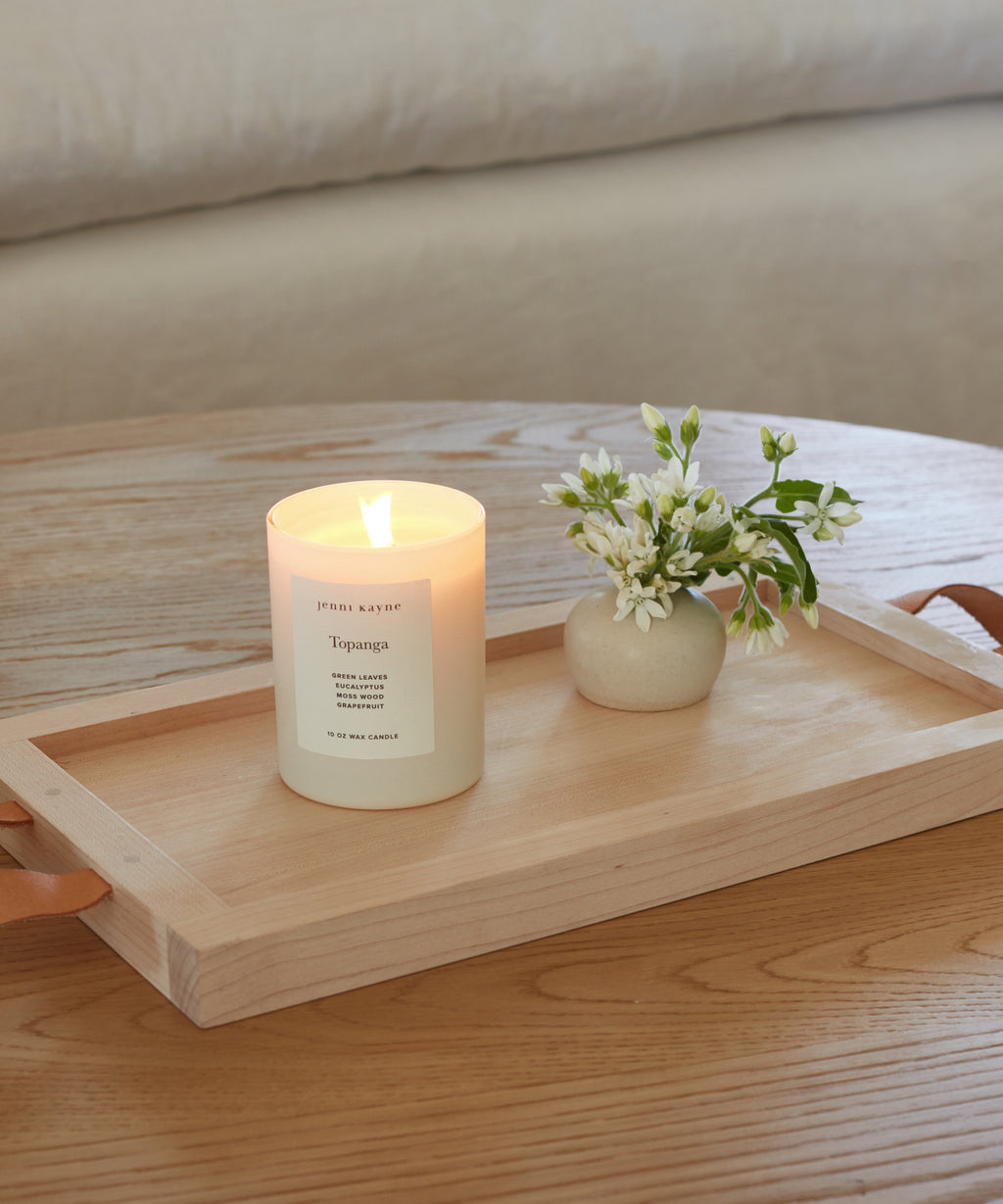 The Topanga Glass Candle, hand-poured in Los Angeles, glows in a glass jar next to white flowers on a light wooden tray with leather handles, adding cozy ambiance to the wooden table.