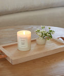 The Topanga Glass Candle, hand-poured in Los Angeles, glows in a glass jar next to white flowers on a light wooden tray with leather handles, adding cozy ambiance to the wooden table.