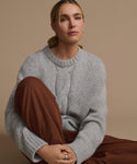 A woman with blonde hair tied back sits against a beige background, wearing the Tove Crewneck in heather grey and loose brown pants. She looks confidently at the camera with a neutral expression.