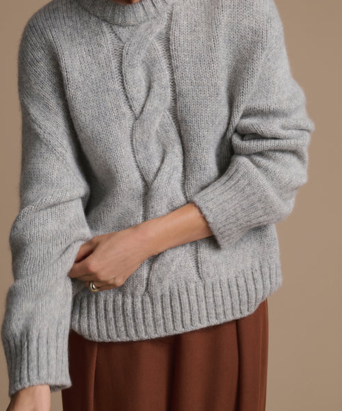 A person wearing the Tove Crewneck, a thick heather grey cable knit sweater with ribbed cuffs and hem, and brown pants stands against a plain beige background. Their head is not visible in the image.