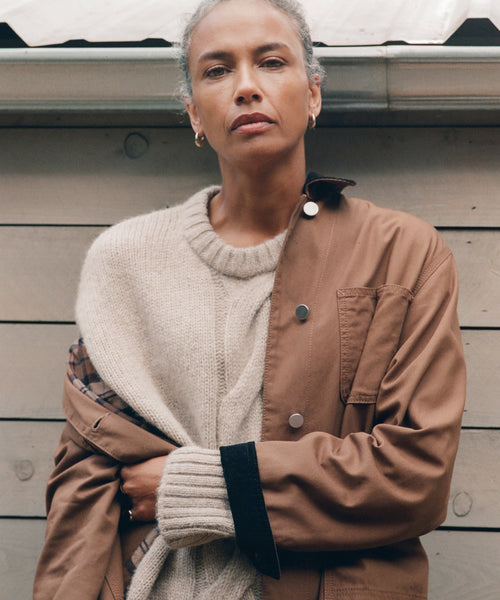 A person with short gray hair stands before a wooden wall, wearing the Tove Crewneck under a brown jacket. They have a neutral expression and look directly at the camera.