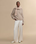 A woman with short hair and small hoop earrings stands against a beige background, wearing the Tove Crewneck sweater with a matching sweater over her shoulders, wide-leg cream satin pants, and cream shoes.