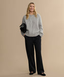 A woman with long wavy blonde hair smiles against a beige background, wearing the loose heather grey Tove Crewneck sweater, wide-leg black pants, black shoes, and holding a small black bag.