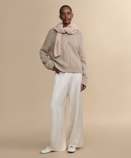 A woman with short hair and small hoop earrings stands against a beige background, wearing the Tove Crewneck sweater with a matching sweater over her shoulders, wide-leg cream satin pants, and cream shoes.
