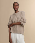 A person with short hair and small earrings stands against a beige background wearing the Tove Crewneck—a loose, chunky cable knit sweater—paired with white pants and their arms crossed.