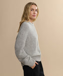 A woman with long blonde hair models the Tove Crewneck, a loose heather grey sweater, paired with black pants. She stands against a beige background, looking confidently at the camera with a slight smile.