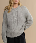 A woman with long blonde hair wears the Tove Crewneck, a heather grey cable knit sweater with a bold front pattern. She stands against a plain beige background, one hand in her pocket.