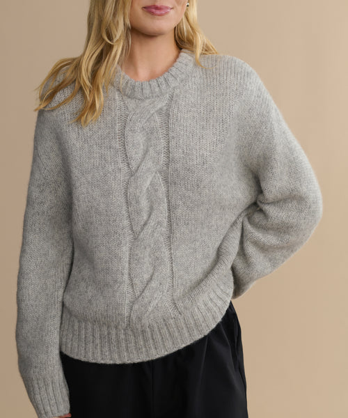 A woman with long blonde hair wears the Tove Crewneck, a heather grey cable knit sweater with a bold front pattern. She stands against a plain beige background, one hand in her pocket.