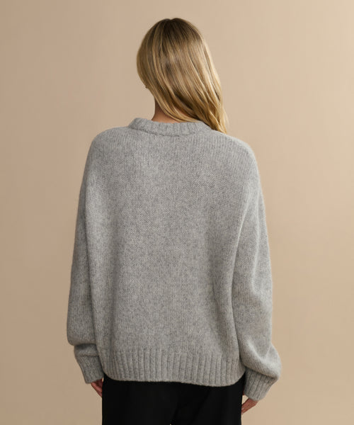 A person with long blonde hair, seen from behind, wears the Tove Crewneck in heather grey with ribbed cuffs and hem, paired with black pants, against a plain beige background.