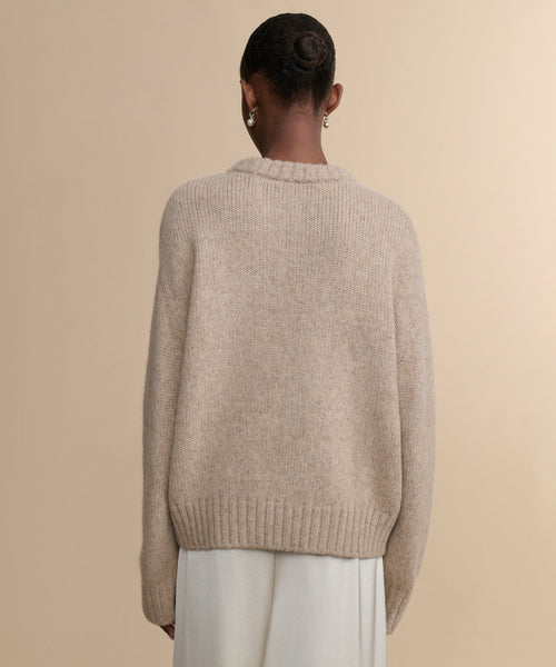A woman with her hair in a bun wears the Tove Crewneck, a chunky beige sweater, paired with a white skirt as she stands facing away from the camera against a plain beige background.