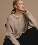 A woman with blonde hair tied back sits on a chair, wearing the Tove Crewneck sweater and black pants, gazing thoughtfully to the side against a plain beige background.