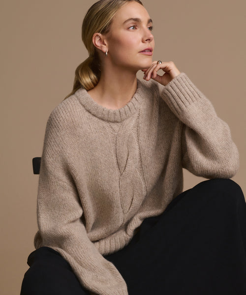 A woman with blonde hair tied back sits on a chair, wearing the Tove Crewneck sweater and black pants, gazing thoughtfully to the side against a plain beige background.