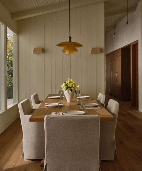 A modern dining room featuring the Trestle Dining Table set for six, white slipcovered chairs, a vase of white lilies centerpiece, pendant lights above, sleek wall sconces, and a large window flooding the space with natural light.