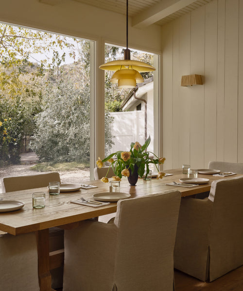 A cozy dining room features the Trestle Dining Table set for six, beige upholstered chairs, a yellow pendant light, and large windows overlooking a sunny garden. Fresh flowers on the table add to the inviting atmosphere.